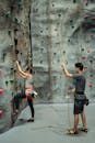 Woman rock climbing indoors with trainer belaying rope for safety.